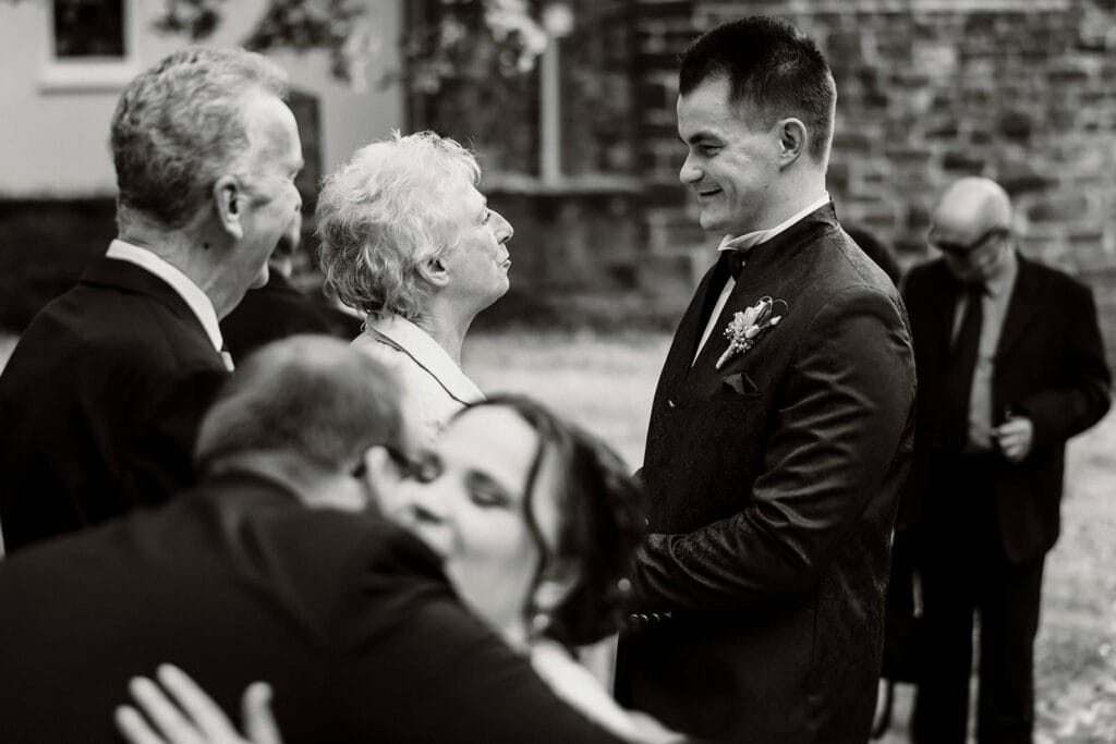 Stiepeler Dorfkirche Landhaus Grum Hattingen Hochzeitsfotograf - Großmutter spricht mit Bräutigam. Im Vordergrund umarmt die Braut den Brautopa