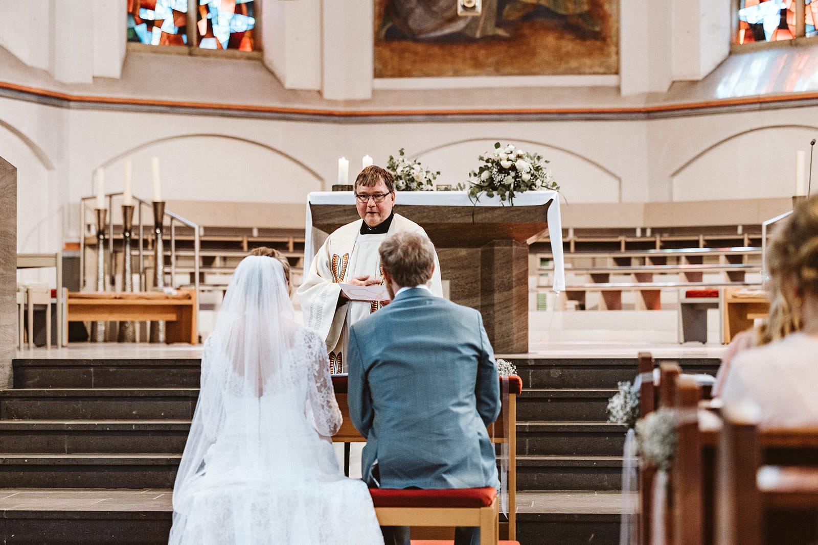 St. Mariä Empfängnis Essen Hochzeitsfotograf - Brautpaar sitzt auf Stühlen vor dem Priester vor dem Altar