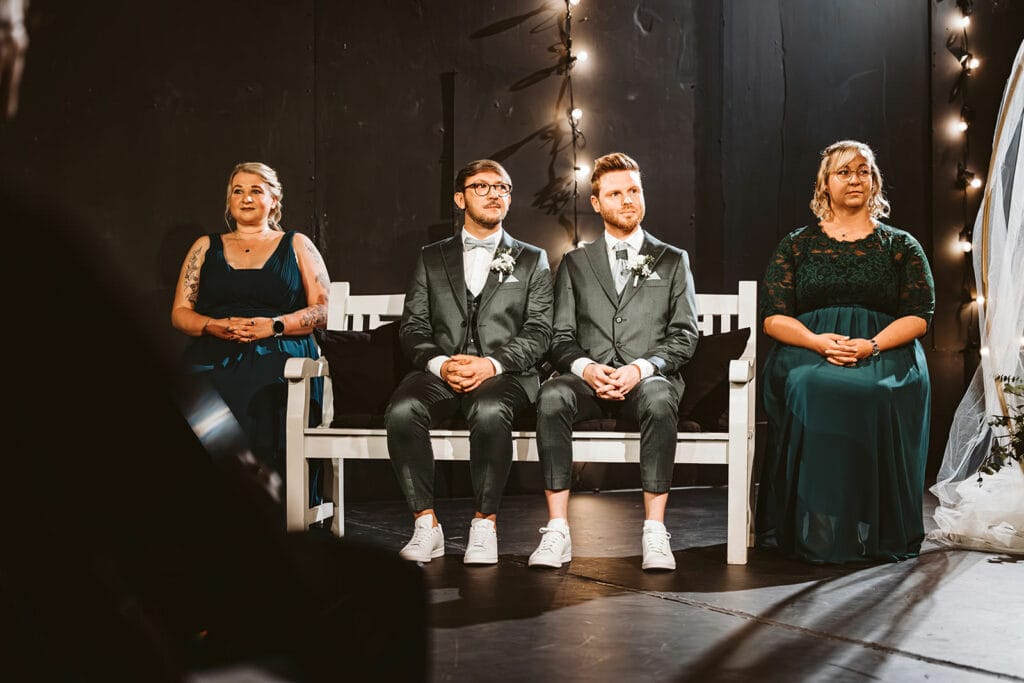 Theater Narrenschiff Unna Lindenbrauerei Kühlschiff Unna Hochzeitsfotograf - Hochzeitspaar Gleichgeschlechtlich Schwul schaut von der Bank aus zur Rednerin