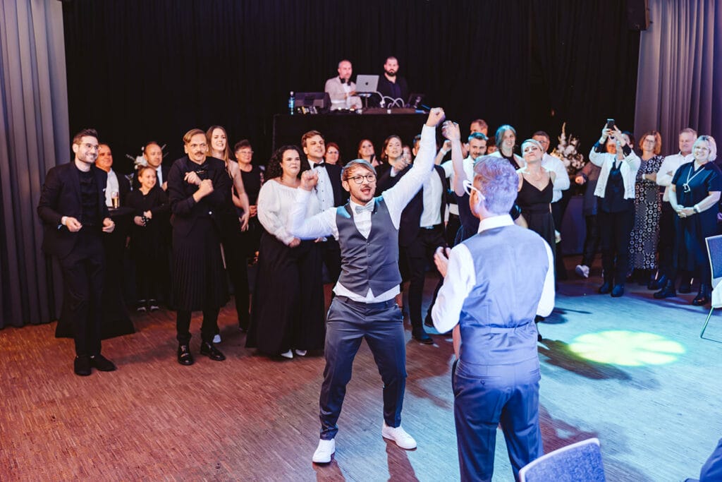 Theater Narrenschiff Unna Lindenbrauerei Kühlschiff Unna Hochzeitsfotograf - Bräutigam hält Choreographie vor schwulen Partner mit Hochzeitsgästen im Hintergrund