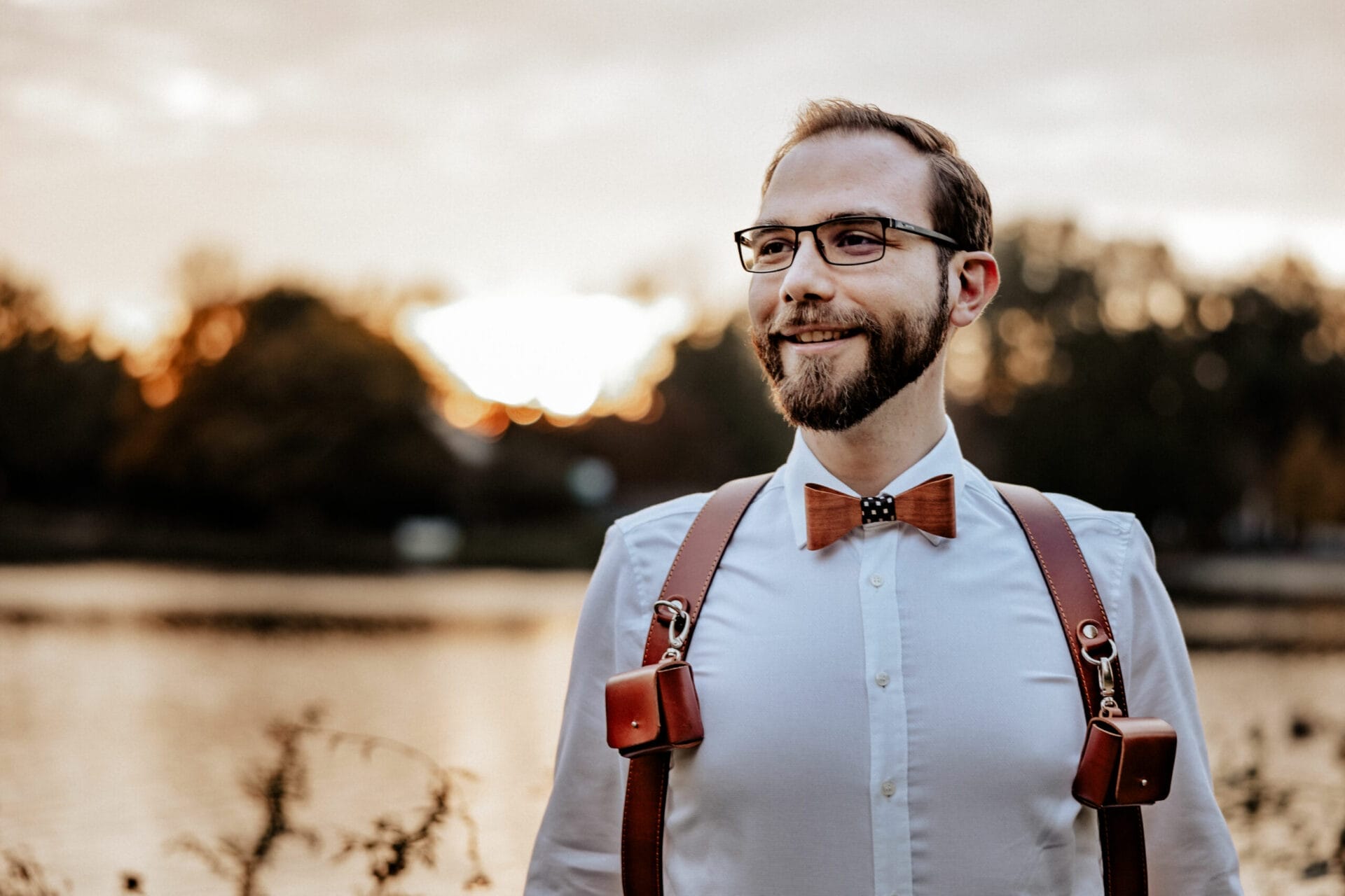 Hochzeit Heiraten Hochzeitsfotograf Dominik Neugebauer - Hochzeitsfotograf mit brauner Fliege und braunem Ledergurt, an dem zwei kleine Taschen aus Leder hängen. Im Hintergrund geht die Sonne unter.