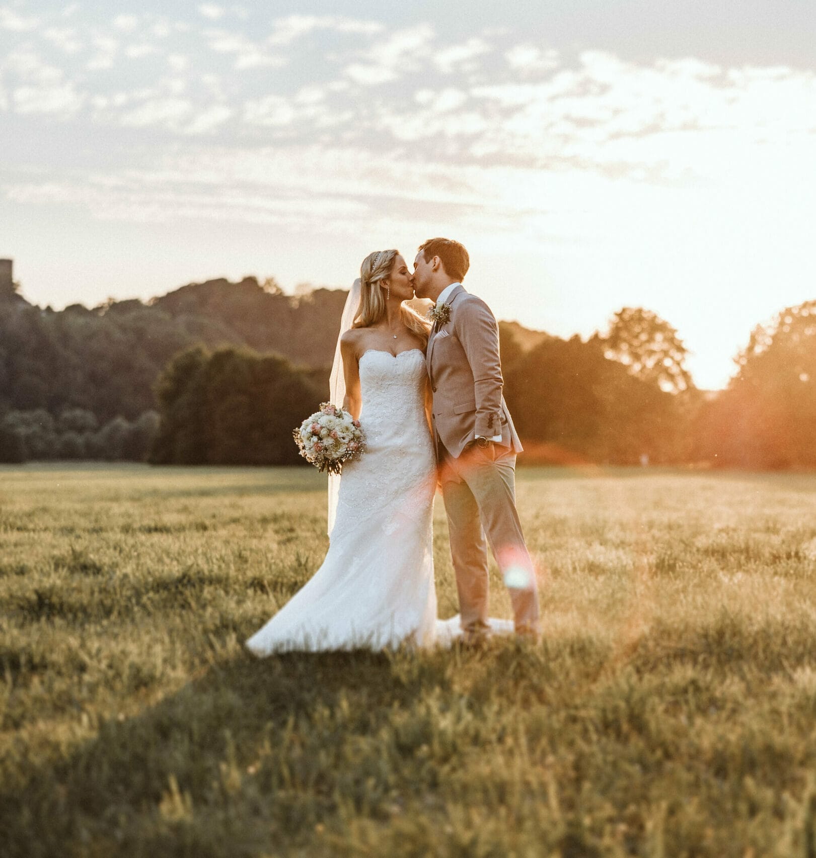 Hochzeit Heiraten Hochzeitsfotograf Dominik Neugebauer - Hochzeitspaar küsst sich auf einer Wiese bei Sonnenuntergang. Bräutigam hat seine linke Hand in der Hosentasche. Die Braut hält ihren Blumenstrauß in der rechten Hand fest. Im Hintergrund ist Wald zu erkennen.