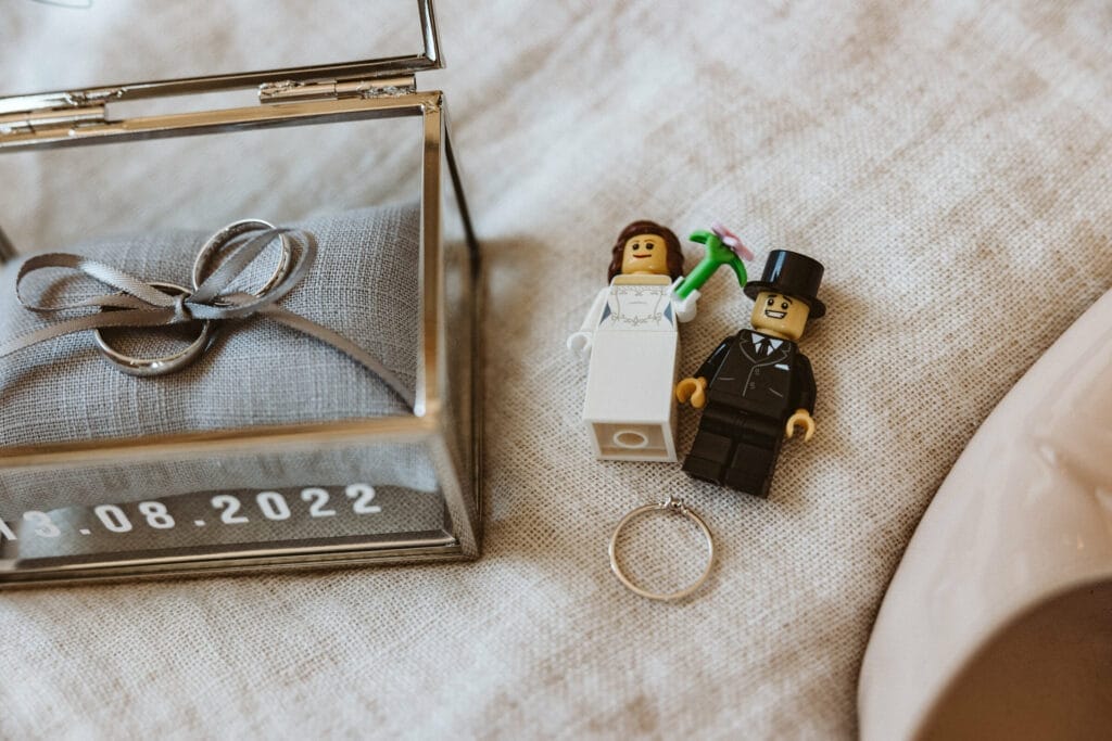 Hochzeit Heiraten Hochzeitsfotograf Dominik Neugebauer - Lego-Figuren von Braut und Bräutigam liegen auf einem Bett. Unter den Figuren befindet sich der Verlobungsringt. Links neben den Lego-Figuren befindet sich ein Schmuck-Kästchen aus Glas. Dort auf einem kleinen Kissen sind die Eheringe mit einem Band verbunden. Auf dem Schmuck-Kästchen steht das Hochzeitsdatum 13.08.2022.