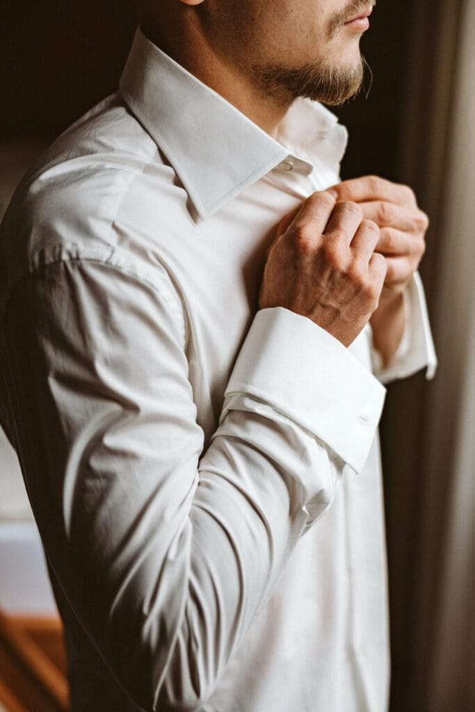 Hochzeit Heiraten Hochzeitsfotograf Dominik Neugebauers - Der Bräutigam zieht sich gerade sein weißes Hemd an. Er knöpft sich den obersten Knopf des Hemds zu und blickt aus dem Fenster.