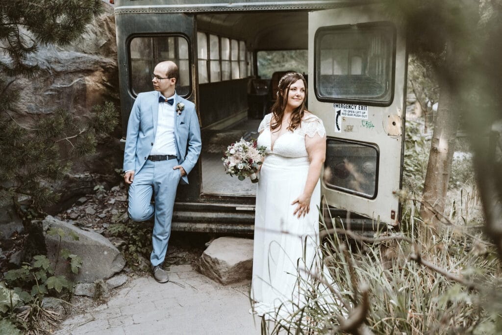 Hochzeit Heiraten Hochzeitsfotograf Dominik Neugebauer - Braut und Bräutigam stehen vor einem alten Schulbus, der die Türen offenstehen hat. Sie schauen in verschiedenen Richtungen. Der Bus steht in dem Tierpark beziehungsweise dem Zoo ZOOM Erlebniswelt.