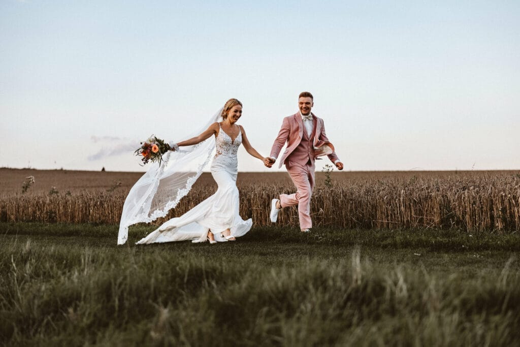 Hochzeit Heiraten Hochzeitsfotograf Dominik Neugebauer - Braut und Bräutigam rennen über die Wiese - Hand in Hand. Hinter ihnen ist das Kornfeld zu sehen. Der Schleier der Braut weht im Wind. Beide lächeln sich bei der Brautpaar-Session verliebt an.
