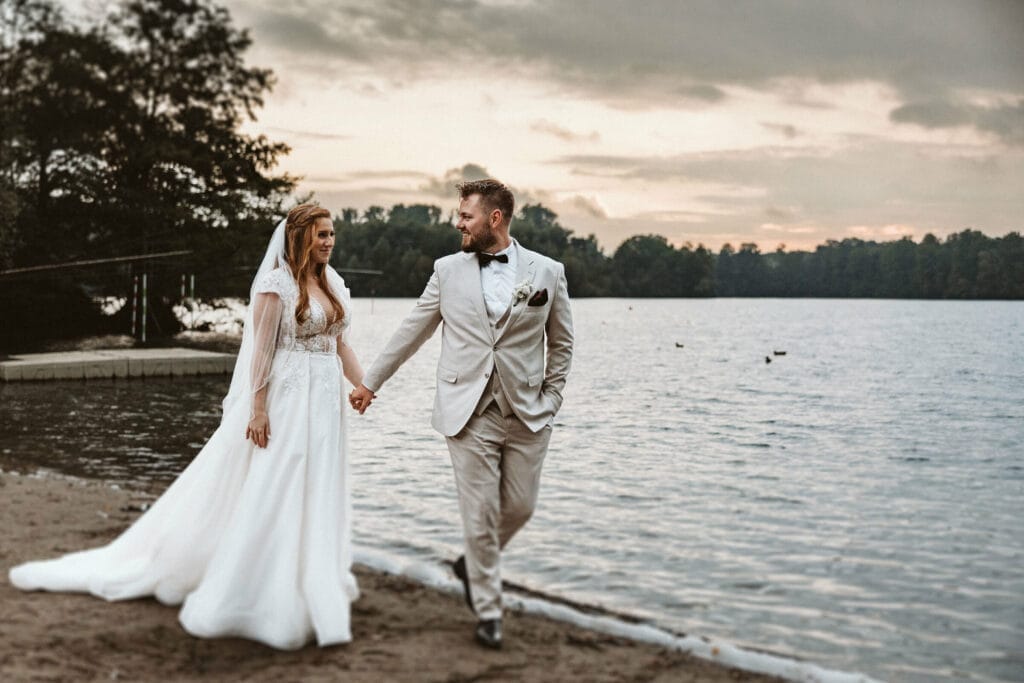 Hochzeit Heiraten Hochzeitsfotograf Dominik Neugebauer - Braut und Bräutigam halten Händchen und laufen am Strand eines Sees entlang. Es ist gerade Sunset und damit Sonnenuntergang. Beide lächeln sich verliebt an.