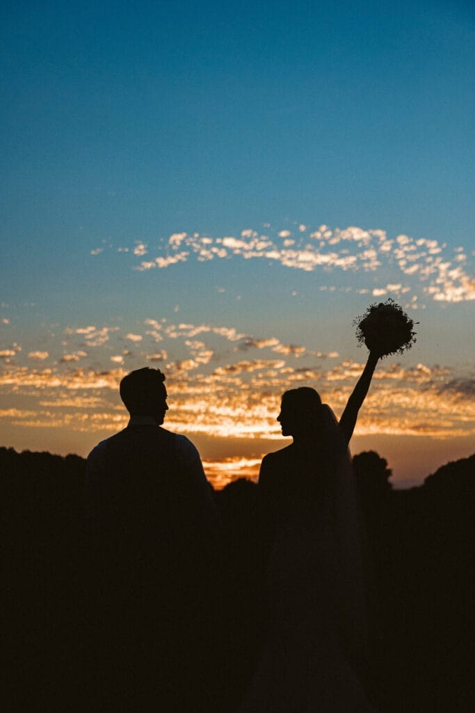 Hochzeit Heiraten Hochzeitsfotograf Dominik Neugebauer - Die Silhouetten der Braut und des Bräutigams sind bei der Sonnenuntergangs-Stimmung zu erkennen. Die Braut schaut zu ihrem Bräutigam und streckt die Hand mit dem Brautstrauß in die Luft.