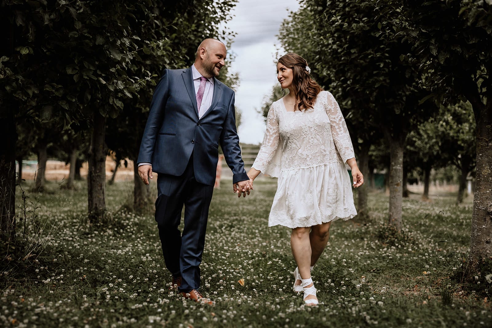 Hochzeit Heiraten Hochzeitsfotograf Dominik Neugebauer Portfolio - Braut läuft mit Bräutigam an Apfelbaum-Allee Hand in Hand. Sie sehen sich beide einander an