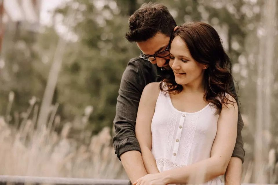 Hochzeitstradition Verlobungsshooting - Engagement Session Hochzeitstraditionen vor dem großen Tag - Pärchen umarmt sich von hinten im Kornfeld und kuschelt miteinander