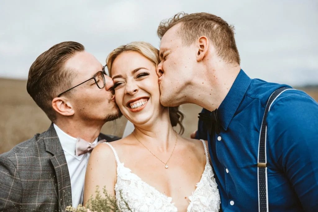 So entstehen tolle Gruppenfotos für die Hochzeit - Braut wird von Trauzeugen auf die Wange geküsst. Die Braut lächelt dabei