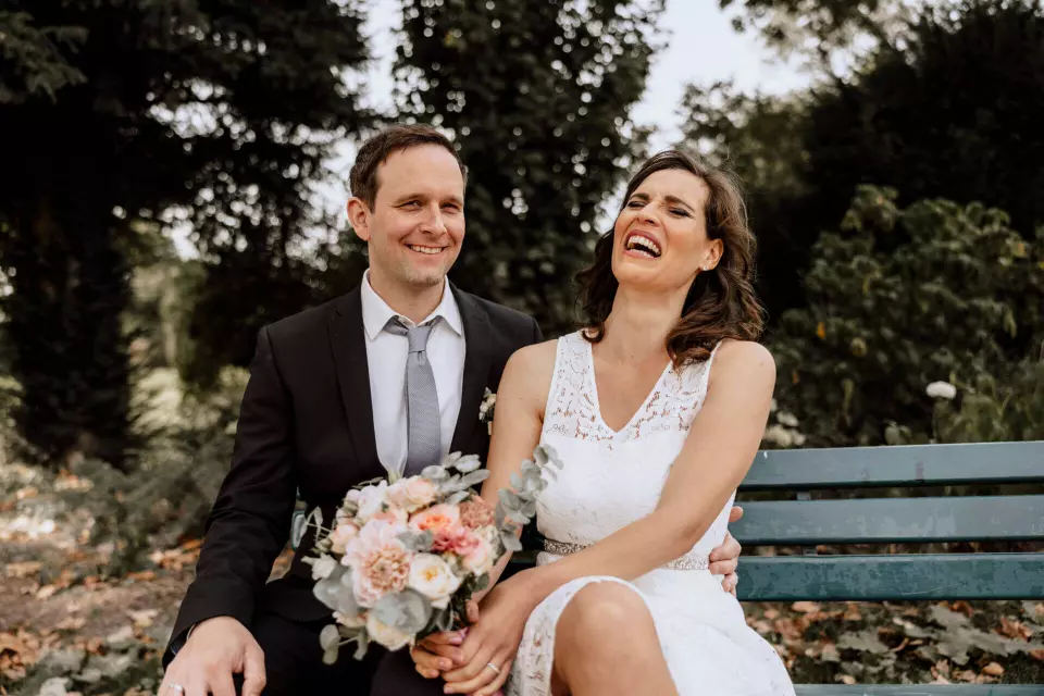 Warum wurde früher in schwarz geheiratet - Braut und Bräutigam sitzen auf einer grünen Bank und lachen. Braut hält Blumenstrauß fest