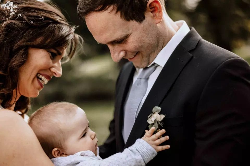 Was kostet ein guter Hochzeitsfotograf - Brautpaar mit Baby im Arm. Baby greift nach Ansteck-Blume des Vaters Bräutigam