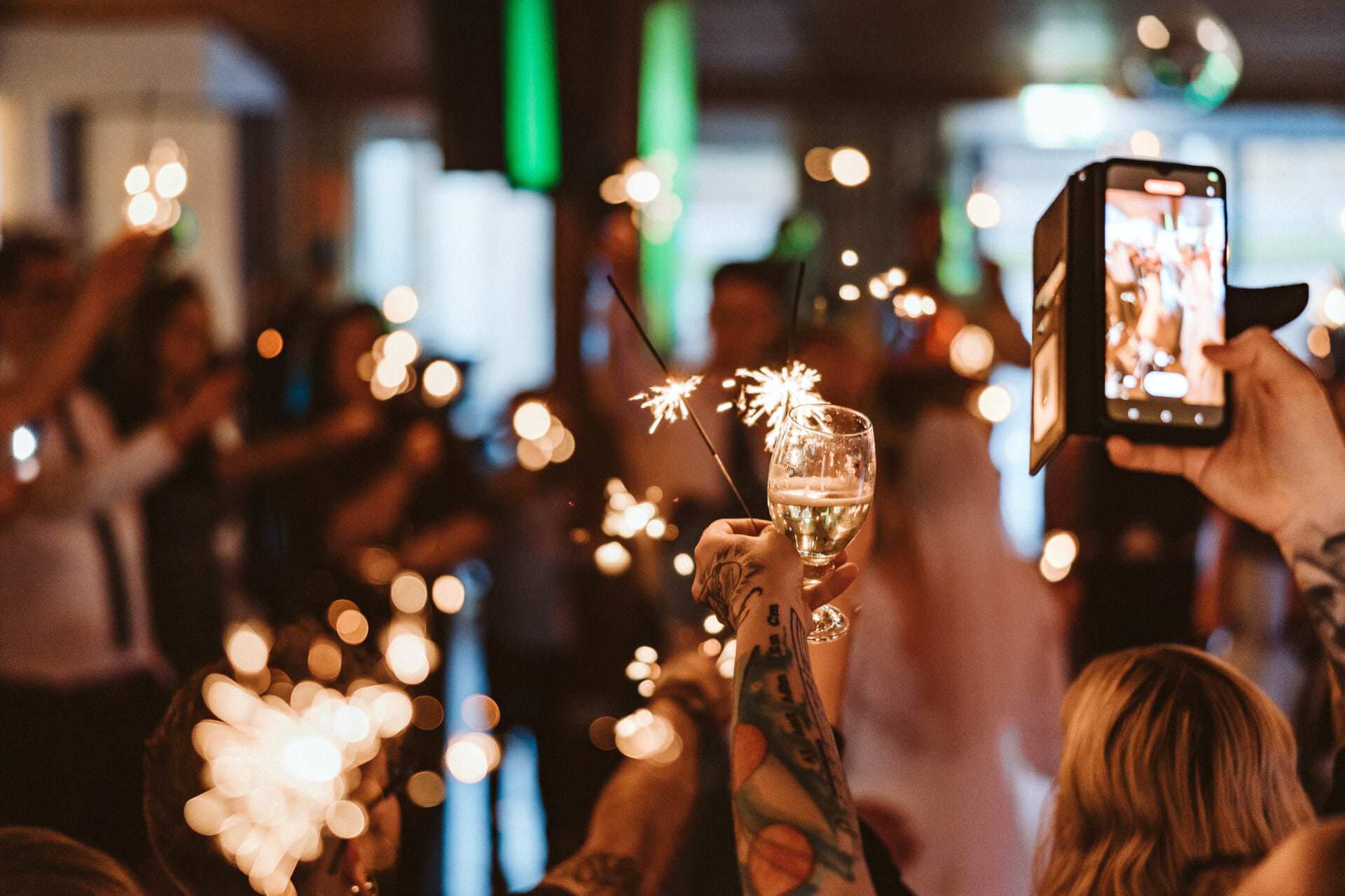 Hochzeitsfotograf Bochum Hochzeit Heiraten Hochzeitsfotos - Beginn des Hochzeitstanzes des Brautpaars mit einem Sektglas in der Hand. Hochzeitsgäste halten Handys in die Luft sowie Wunderkerzen