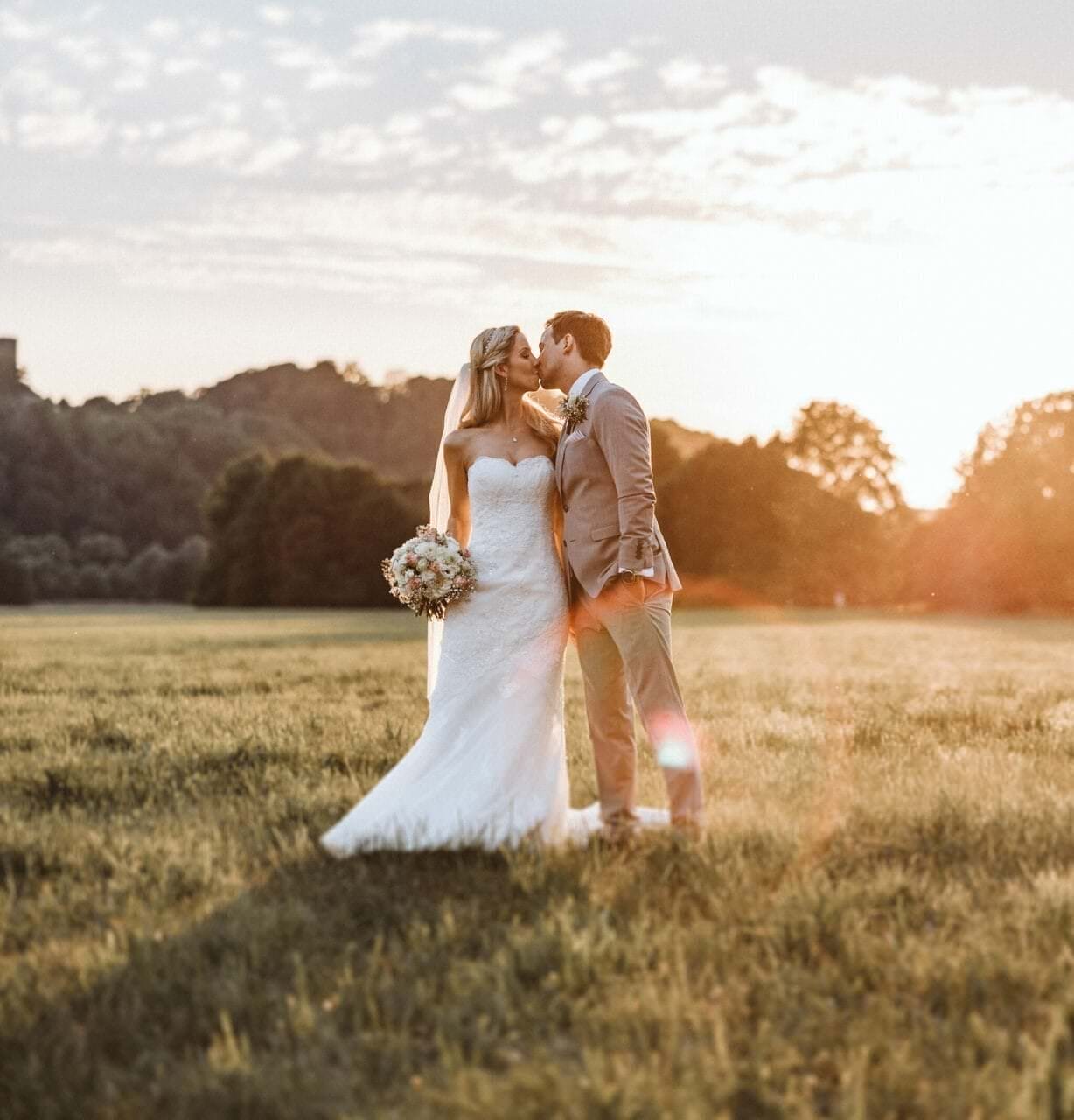 Hochzeitsfotograf Bochum Hochzeit Heiraten Hochzeitsfotos - Hochzeitspaar küsst sich auf einer Wiese bei Sonnenuntergang. Bräutigam hat seine linke Hand in der Hosentasche. Die Braut hält ihren Blumenstrauß in der rechten Hand fest. Im Hintergrund ist Wald zu erkennen