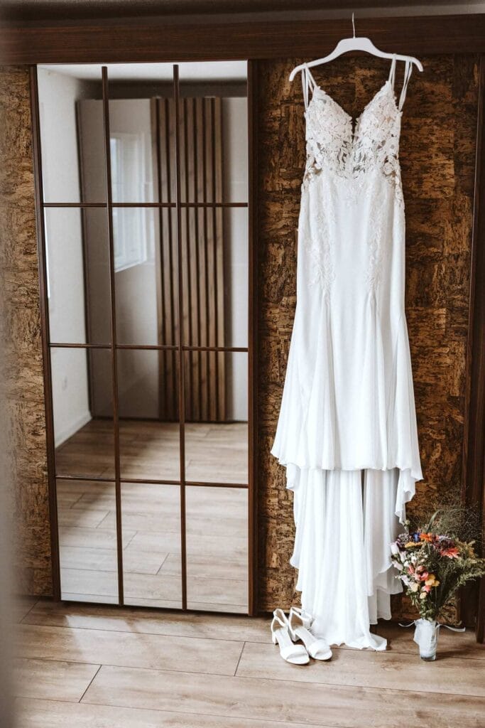 Hochzeitsfotograf Bochum Hochzeit Heiraten Hochzeitsfotos - Brautkleid hängt am weißen Kleiderbügel neben einem Spiegel. Unter dem Brautkleid sind weiße Brautschuhe zu erkennen. Direkt daneben der Brautstrauß der Braut