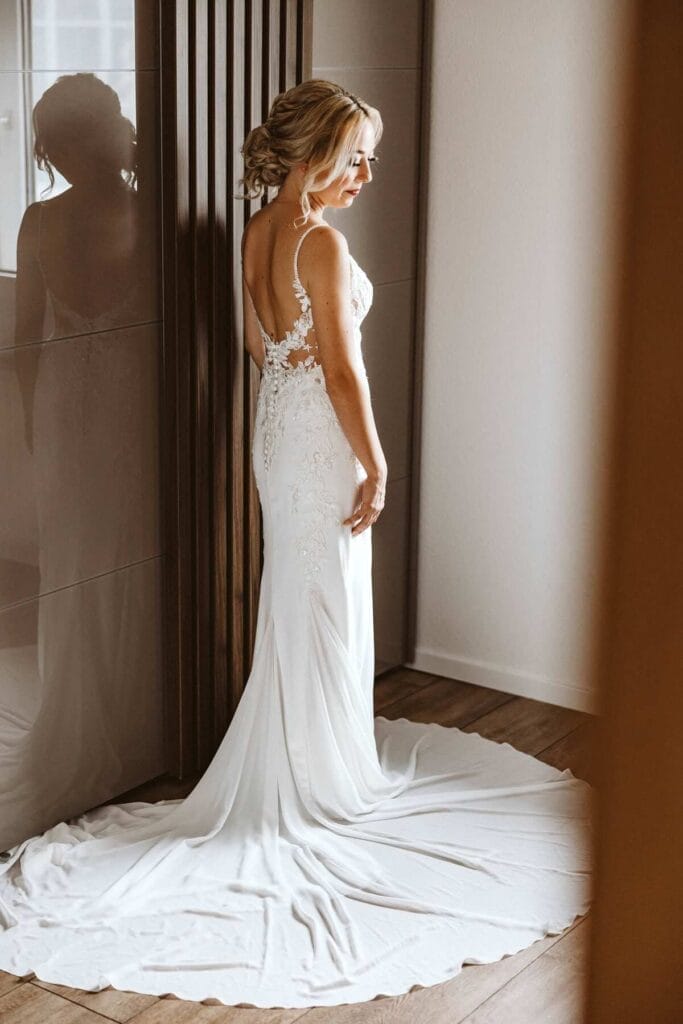 Hochzeitsfotograf Remscheid Hochzeit Heiraten Hochzeitsfotos - Braut streichelt sich leicht mit ihrer rechten Hand an ihre rechte hüfte. Das Brautkleid ist ausgefächert und liegt auf dem Boden. Die Braut schaut leicht schräg nach unten. Im Spiegel, der sich direkt daneben befindet, erkennt man ihre Kontur
