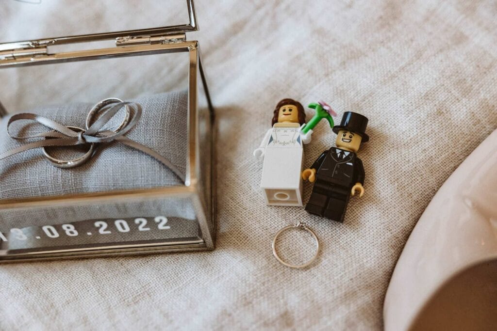 Hochzeitsfotograf Bochum Hochzeit Heiraten Hochzeitsfotos - Lego-Figuren von Braut und Bräutigam liegen auf einem Bett. Unter den Figuren befindet sich der Verlobungsringt. Links neben den Lego-Figuren befindet sich ein Schmuck-Kästchen aus Glas. Dort auf einem kleinen Kissen sind die Eheringe mit einem Band verbunden. Auf dem Schmuck-Kästchen steht das Hochzeitsdatum 13.08.2022