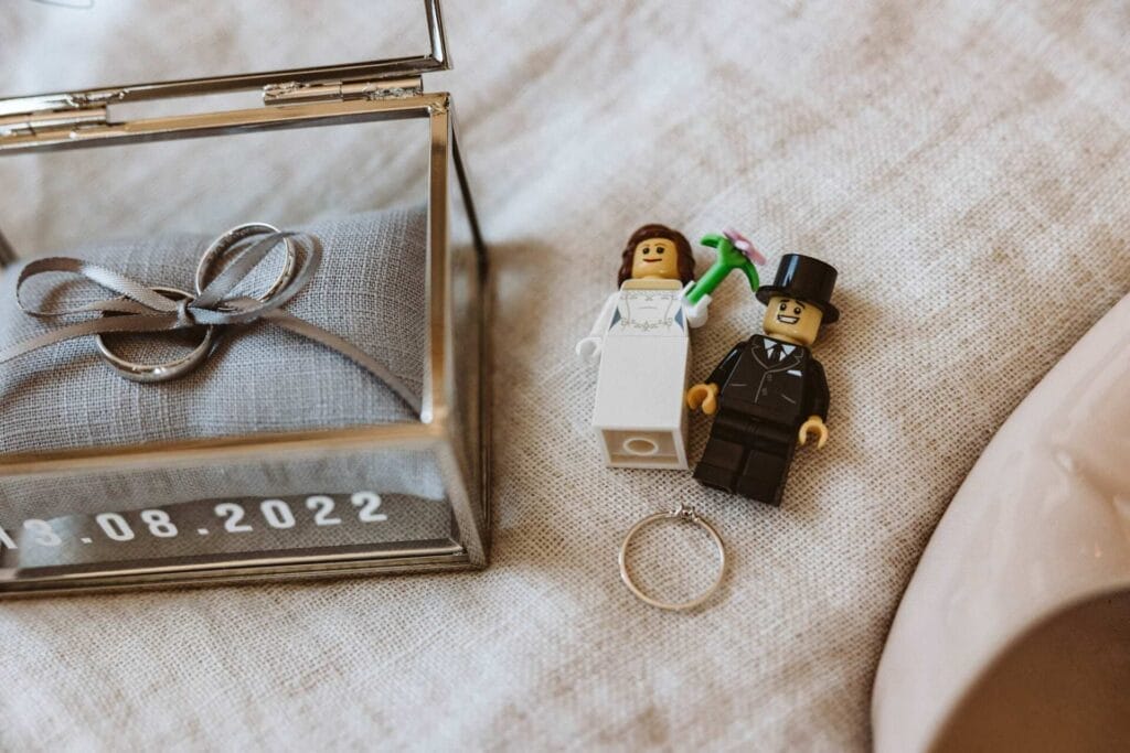 Hochzeitsfotograf Remscheid Hochzeit Heiraten Hochzeitsfotos - Lego-Figuren von Braut und Bräutigam liegen auf einem Bett. Unter den Figuren befindet sich der Verlobungsringt. Links neben den Lego-Figuren befindet sich ein Schmuck-Kästchen aus Glas. Dort auf einem kleinen Kissen sind die Eheringe mit einem Band verbunden. Auf dem Schmuck-Kästchen steht das Hochzeitsdatum 13.08.2022