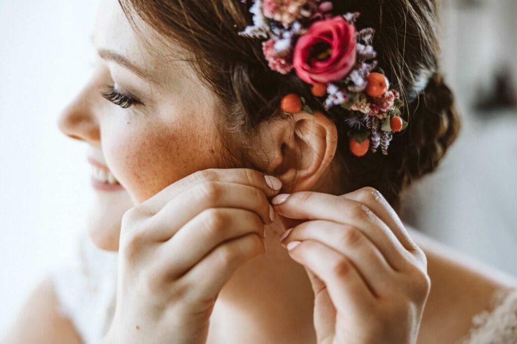 Hochzeitsfotograf Remscheid Hochzeit Heiraten Hochzeitsfotos - Braut mit Blumen im Haar bringt sich einen Ohrschmuck mit beiden Händen an. Der Ohrring ist für die Hochzeit gedacht