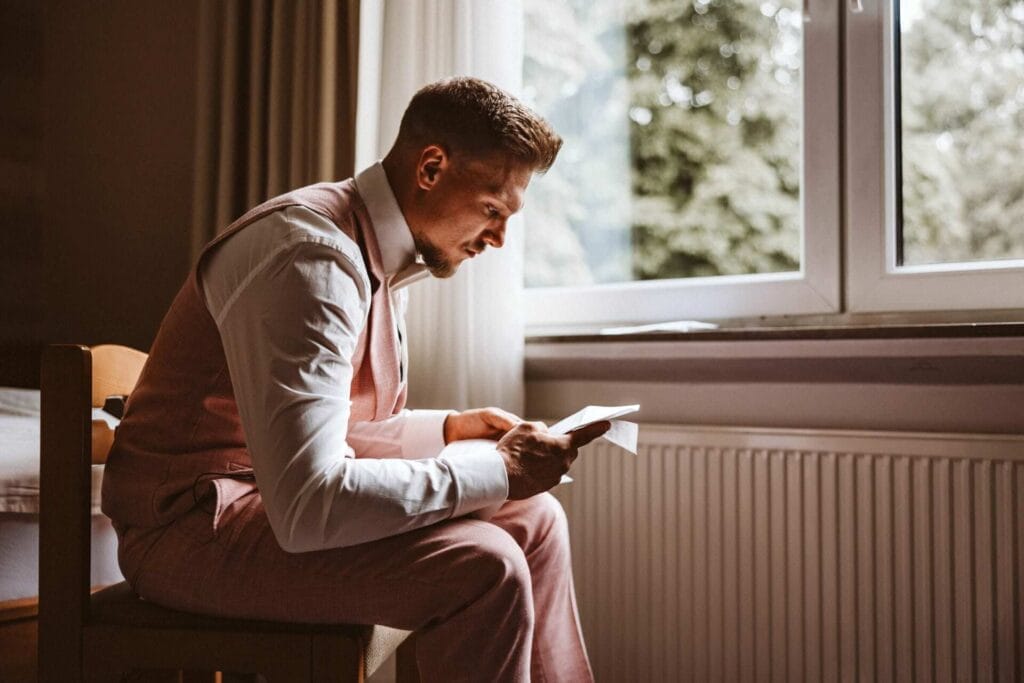 Hochzeitsfotograf Remscheid Hochzeit Heiraten Hochzeitsfotos - Der Bräutigam sitzt auf einem Stuhl. Er hat das Ehegelübde seiner zukünftigen Braut in der Hand und liest sich dieses sorgfältig durch. Er ist dabei den Tränen nahe. Er sitzt vor einem Fenster