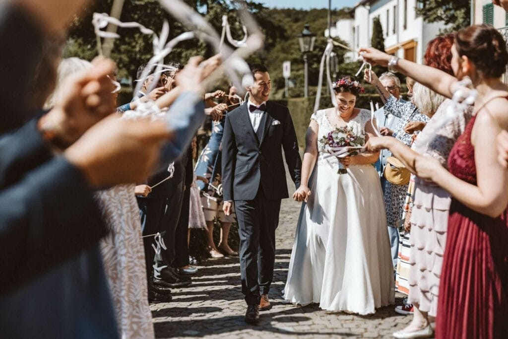 Hochzeitsfotograf Remscheid Hochzeit Heiraten Hochzeitsfotos - Braut und Bräutigam halten ihre Hände und sind überglücklich. Der Bräutigam sieht seine Braut an. Die Braut schaut lächelnd zu Boden. Sie gehen durch ihre Hochzeitsgäste hindurch. Die Hochzeitsgäste bejubeln das Brautpaar und schwingen kleine, selbstgemachte Holzstäbe mit weißen Bändern
