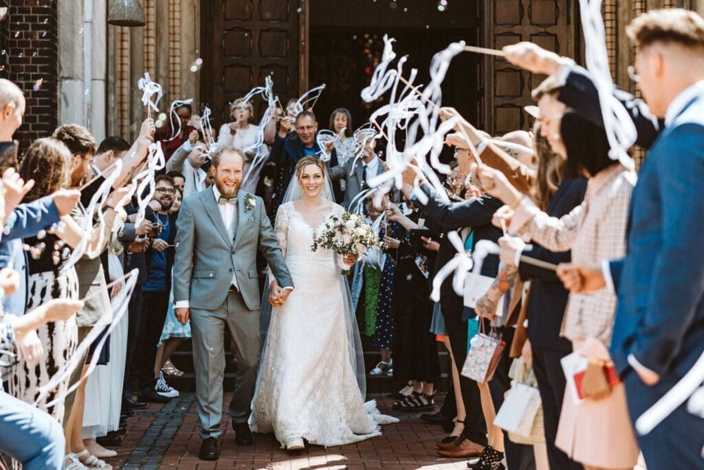 Hochzeitsfotograf Remscheid Hochzeit Heiraten Hochzeitsfotos - Braut und Bräutigam gehen durch ein Spalier von Hochzeitsgästen. Seifenblasen fliegen in der Luft. Die Stimmung ist ausgelassen. Die Hochzeitsgäste wedeln mit einem Holzstab, an dem eine weiße Schlaufe befestigt ist