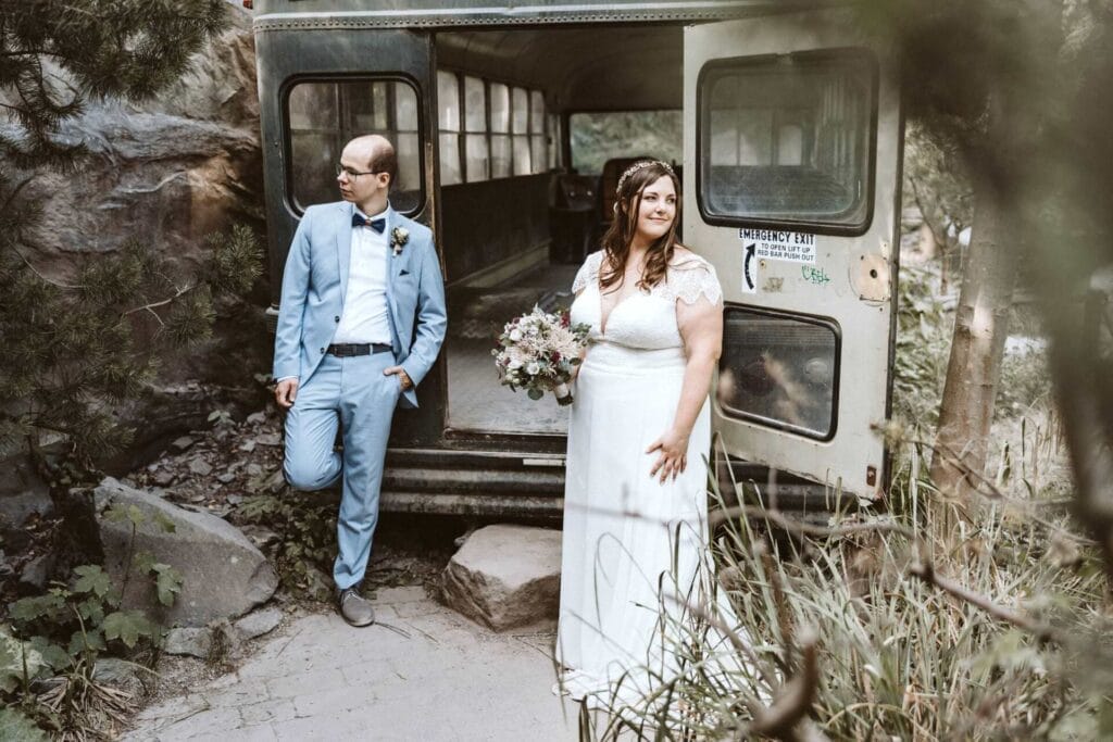 Hochzeitsfotograf Remscheid Hochzeit Heiraten Hochzeitsfotos - Braut und Bräutigam stehen vor einem alten Schulbus, der die Türen offenstehen hat. Sie schauen in verschiedenen Richtungen. Der Bus steht in dem Tierpark beziehungsweise dem Zoo ZOOM Erlebniswelt