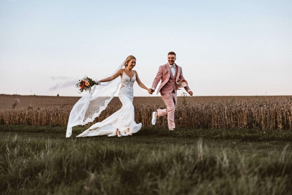 Hochzeitsfotograf Remscheid Hochzeit Heiraten Hochzeitsfotos - Braut und Bräutigam rennen über die Wiese - Hand in Hand. Hinter ihnen ist das Kornfeld zu sehen. Der Schleier der Braut weht im Wind. Beide lächeln sich bei der Brautpaar-Session verliebt an