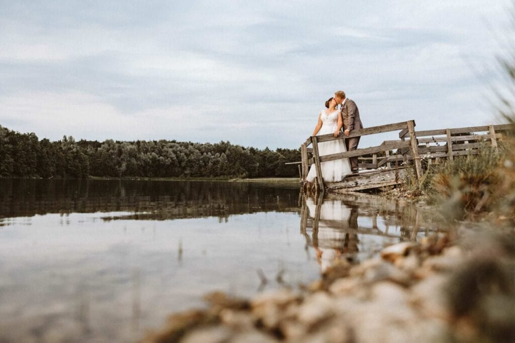 Hochzeitsfotograf Remscheid Hochzeit Heiraten Hochzeitsfotos - Das Hochzeitspaar steht küssend auf einem Steg, welcher ins Wasser führt. Der glasklare See spiegelt das Brautpaar im Wasser