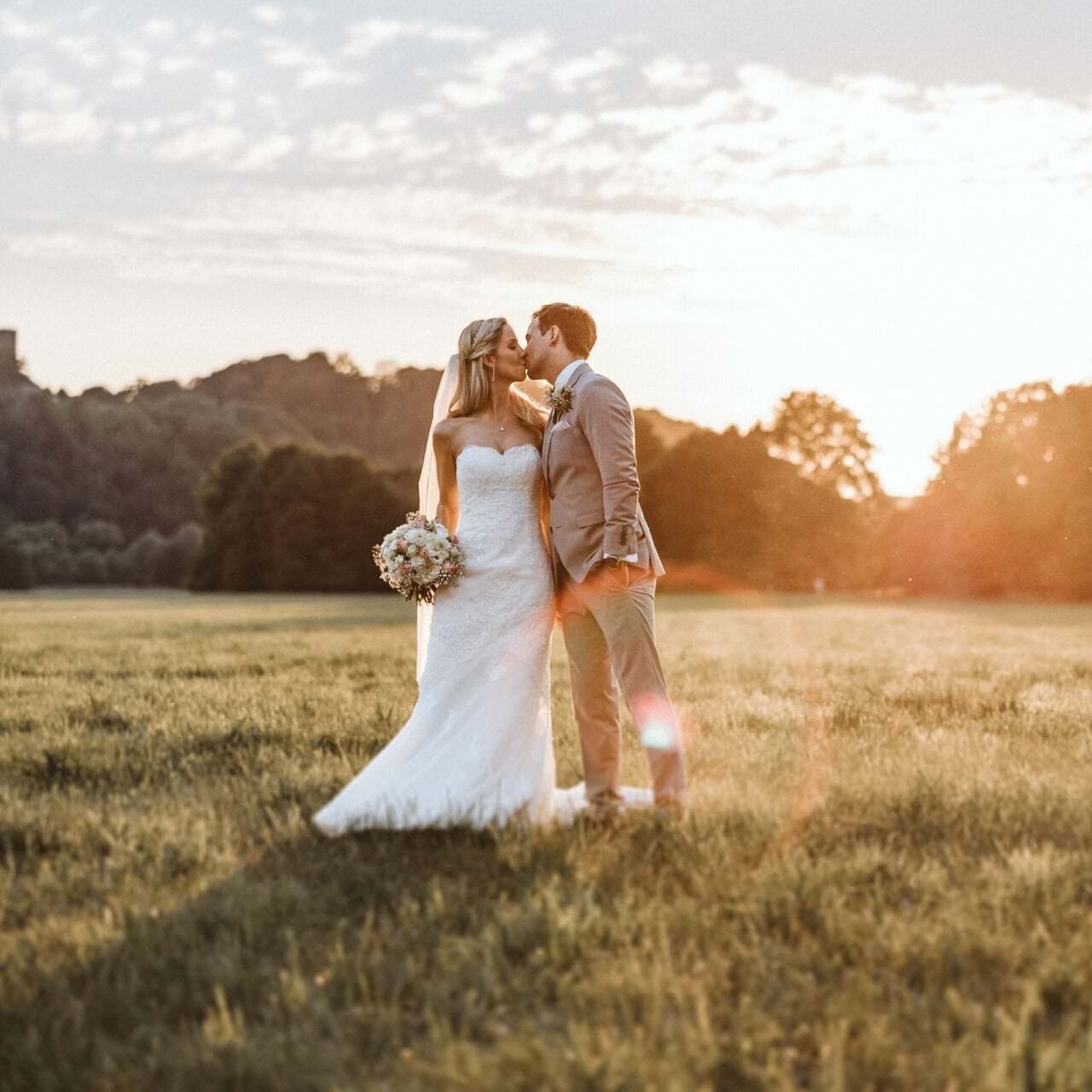 Hochzeitsfotograf Dinslaken Hochzeit Heiraten Hochzeitsfotos - Hochzeitspaar küsst sich auf einer Wiese bei Sonnenuntergang. Bräutigam hat seine linke Hand in der Hosentasche. Die Braut hält ihren Blumenstrauß in der rechten Hand fest. Im Hintergrund ist Wald zu erkennen
