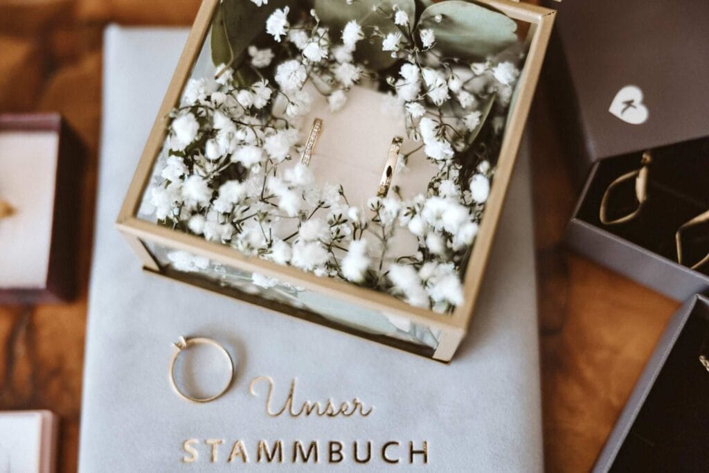 Hochzeitsfotograf Duisburg Hochzeit Heiraten Hochzeitsfotos - Diverser Schmuck, wie der Verlobungsring, die Hochzeits-Ringe sowie ein Stammbuch des Hochzeitspaars liegt neben Ohrringen auf einem Tisch dekoriert. Schleierkraut hübschen die Szene auf