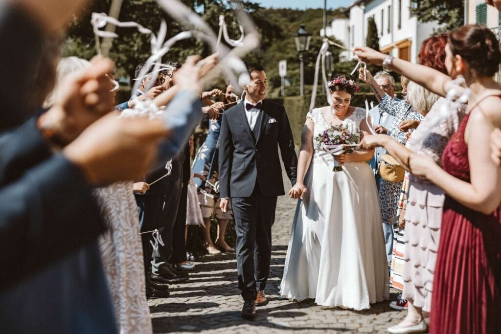 Hochzeitsfotograf Duisburg Hochzeit Heiraten Hochzeitsfotos - Braut und Bräutigam halten ihre Hände und sind überglücklich. Der Bräutigam sieht seine Braut an. Die Braut schaut lächelnd zu Boden. Sie gehen durch ihre Hochzeitsgäste hindurch. Die Hochzeitsgäste bejubeln das Brautpaar und schwingen kleine, selbstgemachte Holzstäbe mit weißen Bändern