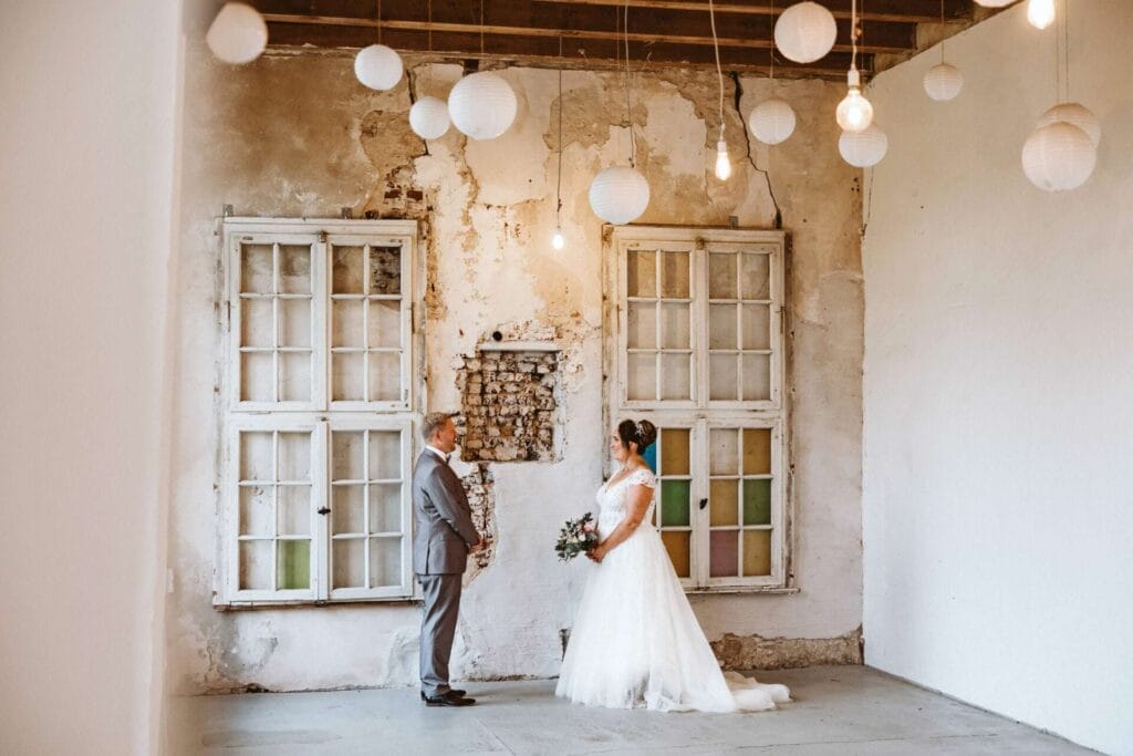 Hochzeitsfotograf Mülheim an der Ruhr Hochzeit Heiraten Hochzeitsfotos - Braut und Bräutigam sind in einem alten Raum, dessen Wände weiß gestrichen sind. Hinter dem Brautpaar sind zwei alte Fenster, die mit verschiedenen Glasfarben verziert sind. Von oben herab hängen kleine Lampen in Kugelform bei der Brautpaar-Session