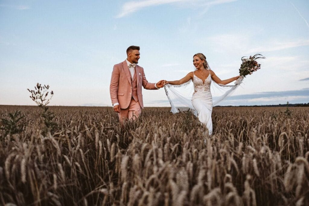 Hochzeitsfotograf Münster Münsterland Hochzeit Heiraten Hochzeitsfotos - Hochzeitspaar läuft Hand in Hand durchs Kornfeld. Beide lächeln sich an. Die Braut hält ihren Blumenstrauß in die Luft