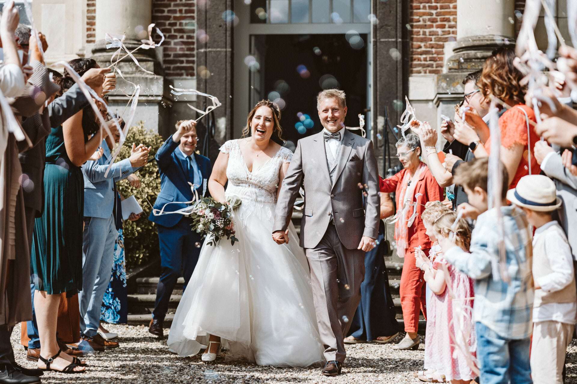 Hochzeitsfotograf Oberhausen Hochzeit Heiraten Hochzeitsfotos - Hochzeitspaar geht Hand in Hand am Spalier der Hochzeitsgäste vorbei. Hochzeitsauszug mit bunten Seifenblasen, Kindern, die Fähnchen schwingen sowie lachende Gäste sind zu erkennen