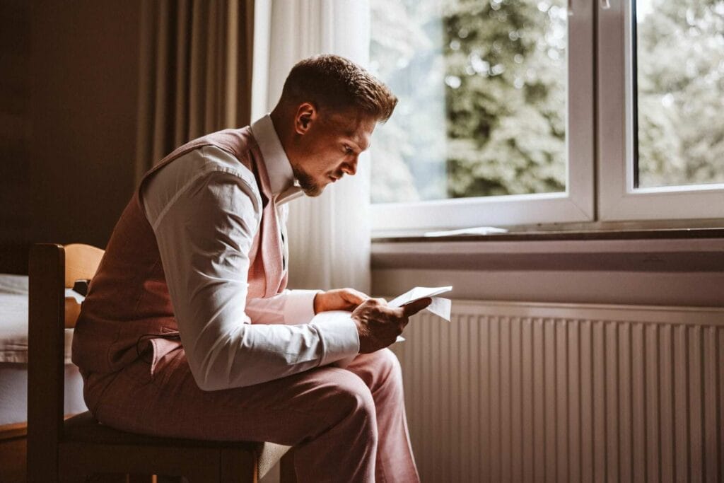 Hochzeitsfotograf Oberhausen Hochzeit Heiraten Hochzeitsfotos - Der Bräutigam sitzt auf einem Stuhl. Er hat das Ehegelübde seiner zukünftigen Braut in der Hand und liest sich dieses sorgfältig durch. Er ist dabei den Tränen nahe. Er sitzt vor einem Fenster