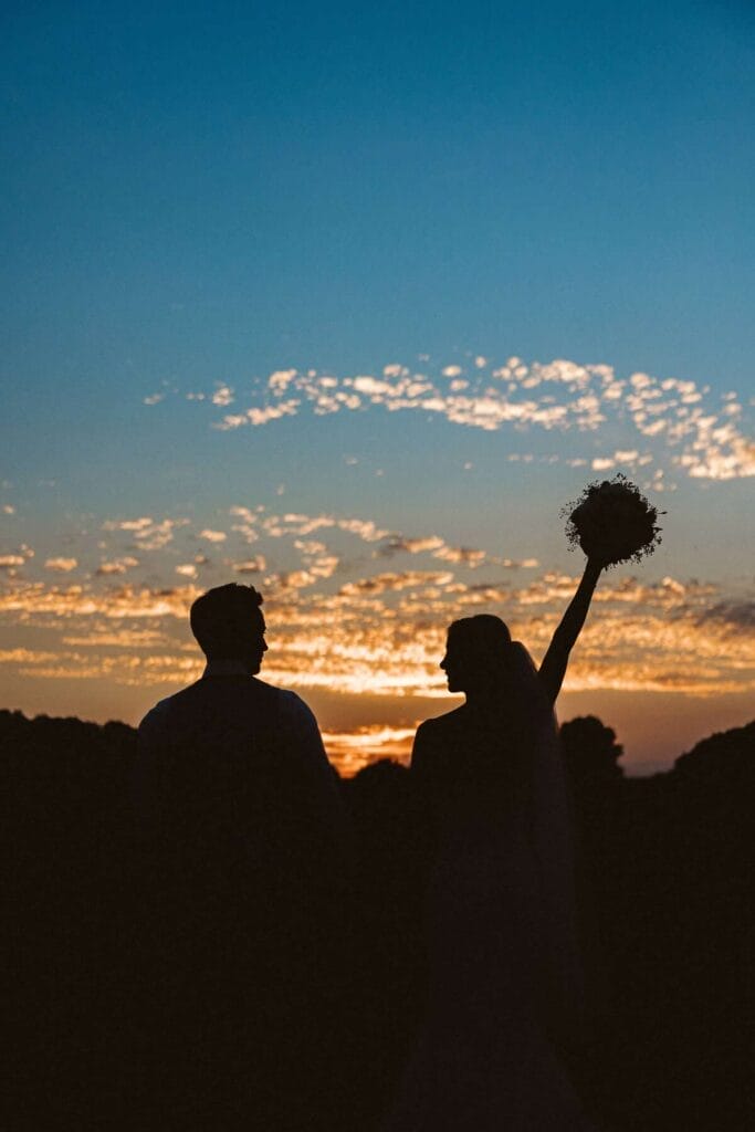 Hochzeitsfotograf Oberhausen Hochzeit Heiraten Hochzeitsfotos - Die Silhouetten der Braut und des Bräutigams sind bei der Sonnenuntergangs-Stimmung zu erkennen. Die Braut schaut zu ihrem Bräutigam und streckt die Hand mit dem Brautstrauß in die Luft