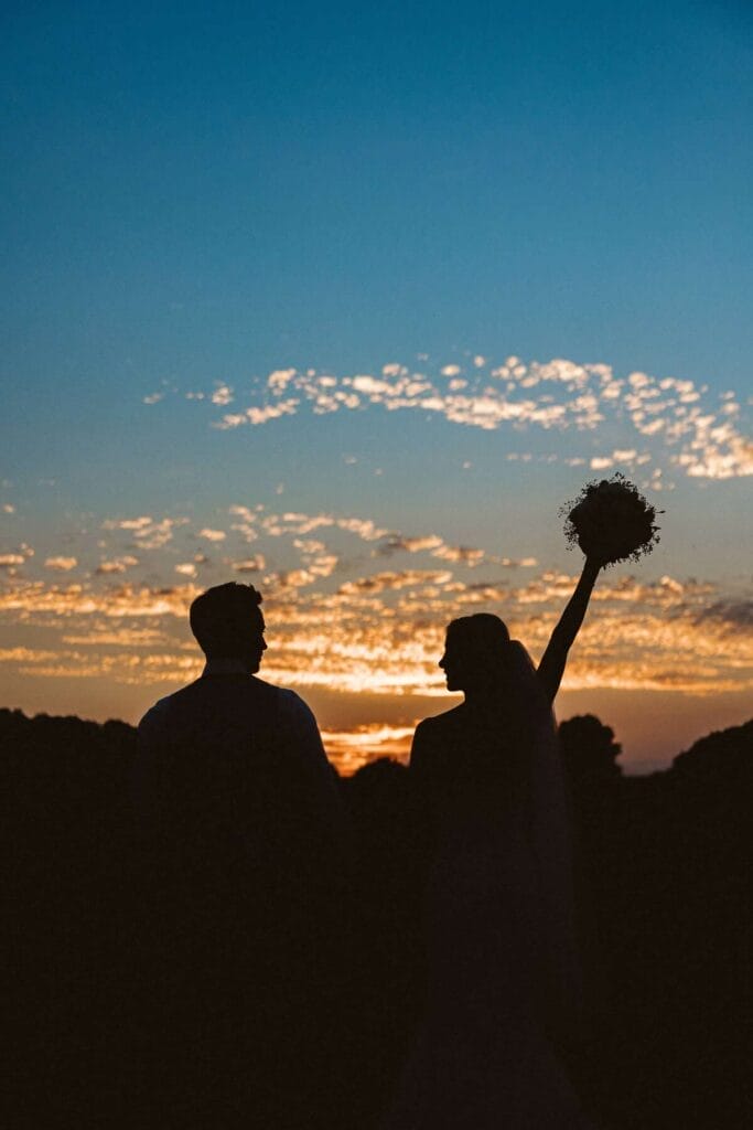 Hochzeitsfotograf Recklinghausen Hochzeit Heiraten Hochzeitsfotos - Die Silhouetten der Braut und des Bräutigams sind bei der Sonnenuntergangs-Stimmung zu erkennen. Die Braut schaut zu ihrem Bräutigam und streckt die Hand mit dem Brautstrauß in die Luft