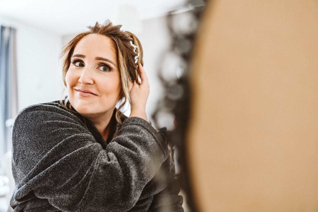 Hochzeitsfotograf Bottrop Hochzeit Heiraten Hochzeitsfotos - Die zukünftige Braut ist in einem schwarzen Kimono gehüllt. Sie überprüft ihre Frisur auf passenden Halt vor einem großen, runden Spiegel, der auf einem Tisch steht. Die zukünftige Braut sieht zufrieden und glücklich aus