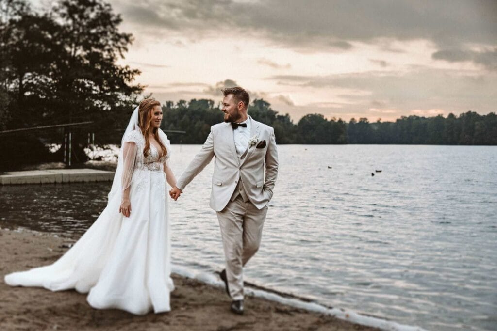 Hochzeitsfotograf Castrop Rauxel Hochzeit Heiraten Hochzeitsfotos - Braut und Bräutigam halten Händchen und laufen am Strand eines Sees entlang. Es ist gerade Sunset und damit Sonnenuntergang. Beide lächeln sich verliebt an