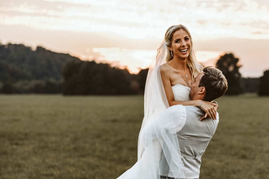 Hochzeitsfotograf Dortmund Hochzeit Heiraten Hochzeitsfotos - Der Bräutigam hebt seine Braut in die Luft und dreht sich mit ihr im Kreis. Die Braut lacht dabei auf und schaut direkt in die Kamera. Im Hintergrund ist der Sonnenuntergang zu sehen. Sie befinden sich auf einem grünen Feld mit Wiese