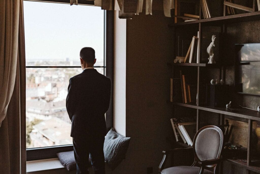 Hochzeitsfotograf Marl Hochzeit Heiraten Hochzeitsfotos - Bräutigam steht vor einem großen Fenster und wirft einen Blick aus einem Hochhaus im Hotel hinaus. Um ihn herum sind Bücher, ein Stuchl sowie ein Fernseher zu erkennen sowie Briefe und Zettel, die aufgehangen sind