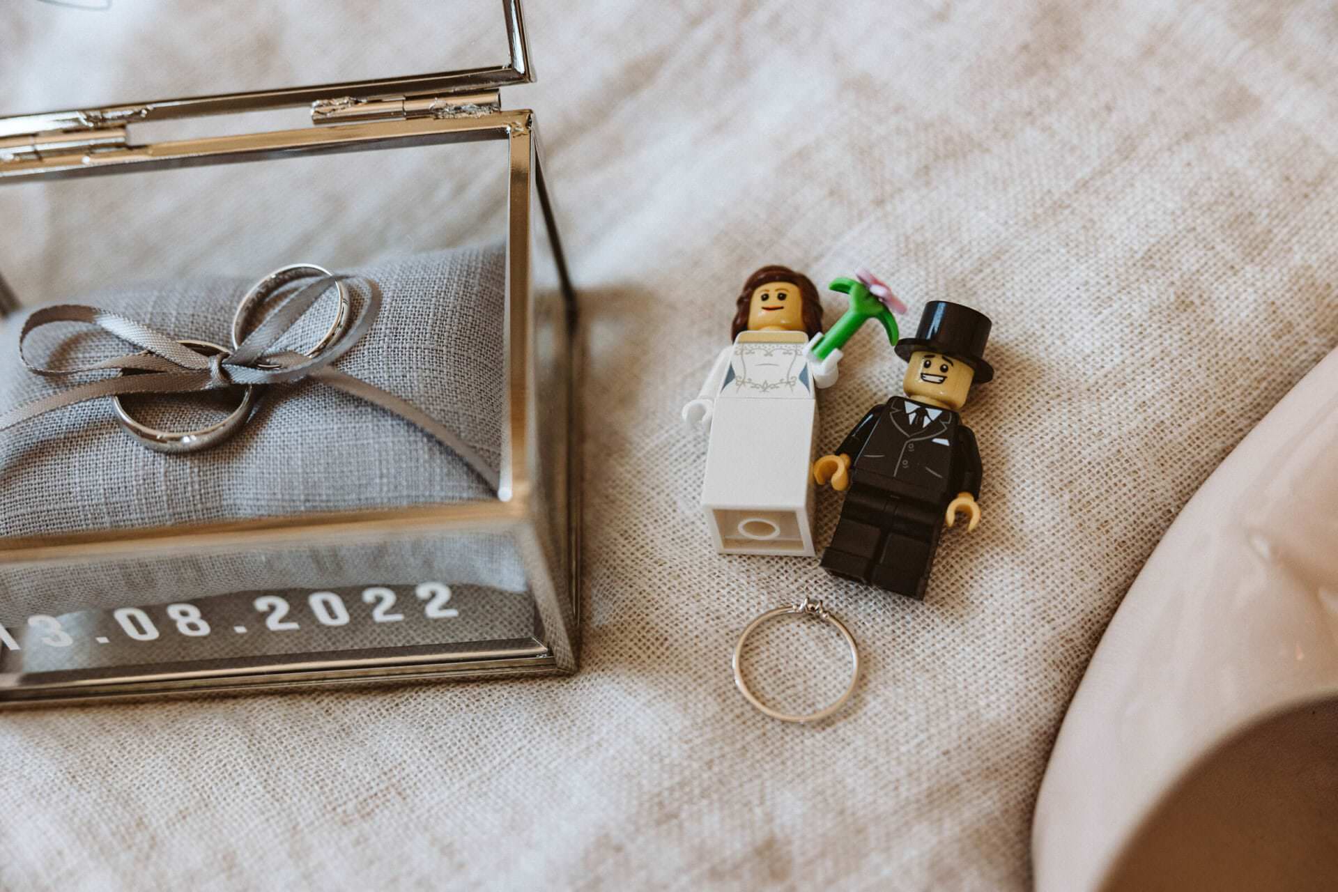 Hochzeitsfotograf Krefeld Hochzeit Heiraten Hochzeitsfotos - Lego-Figuren von Braut und Bräutigam liegen auf einem Bett. Unter den Figuren befindet sich der Verlobungsringt. Links neben den Lego-Figuren befindet sich ein Schmuck-Kästchen aus Glas. Dort auf einem kleinen Kissen sind die Eheringe mit einem Band verbunden. Auf dem Schmuck-Kästchen steht das Hochzeitsdatum 13.08.2022