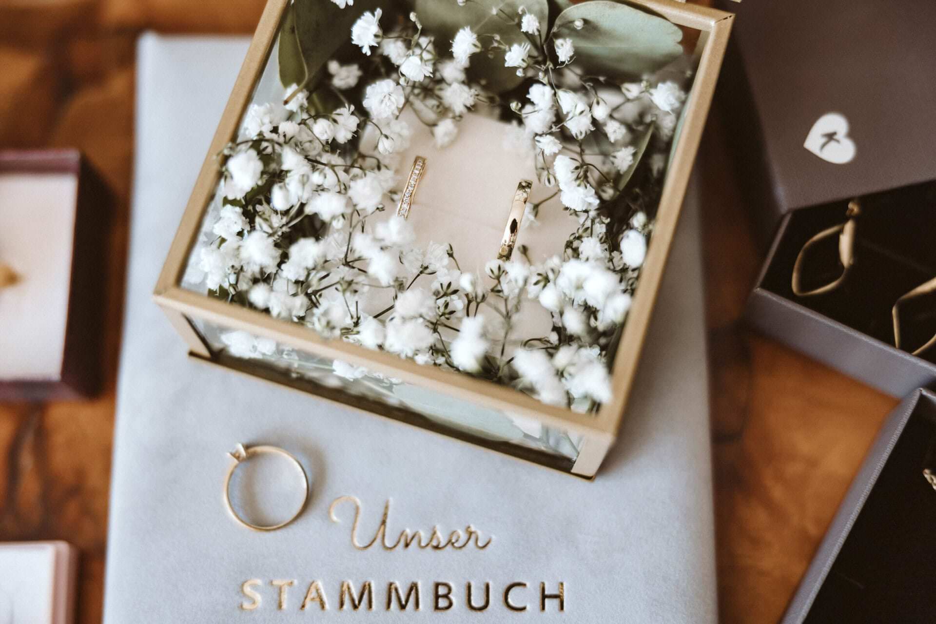 Hochzeitsfotograf Krefeld Hochzeit Heiraten Hochzeitsfotos - Diverser Schmuck, wie der Verlobungsring, die Hochzeits-Ringe sowie ein Stammbuch des Hochzeitspaars liegt neben Ohrringen auf einem Tisch dekoriert. Schleierkraut hübschen die Szene auf