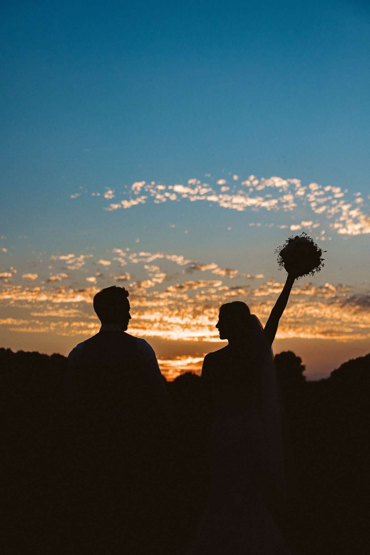 Hochzeitsfotograf Krefeld Hochzeit Heiraten Hochzeitsfotos - Die Silhouetten der Braut und des Bräutigams sind bei der Sonnenuntergangs-Stimmung zu erkennen. Die Braut schaut zu ihrem Bräutigam und streckt die Hand mit dem Brautstrauß in die Luft