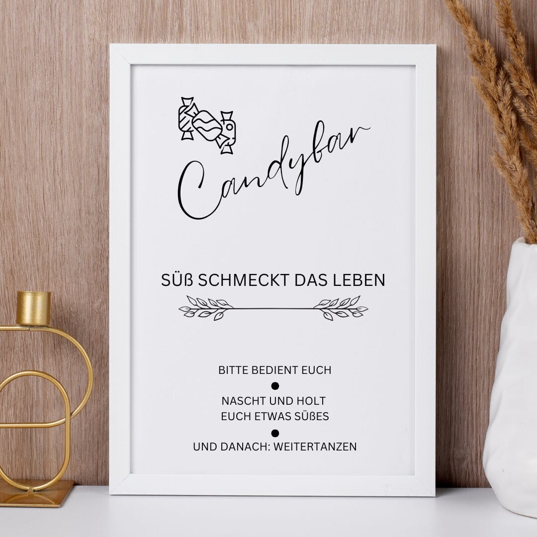 Auf einem gerahmten Schild auf einer hölzernen Fläche steht Candybar in Schreibschrift. Darunter steht auf Deutsch: Süß schmeckt das Leben, gefolgt von der Aufforderung an die Gäste, sich an den Süßigkeiten zu bedienen und weiterzutanzen. Neben dem Rahmen befinden sich dekorative Gegenstände.