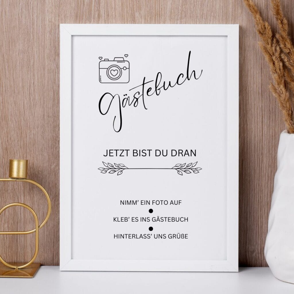 Ein gerahmtes Schild mit deutschem Text und einem Kamerasymbol auf einem hölzernen Hintergrund, neben einem goldenen Kerzenhalter und getrocknetem Gras. Das Schild lädt die Gäste ein, ein Foto und eine Nachricht in ein Gästebuch einzutragen.