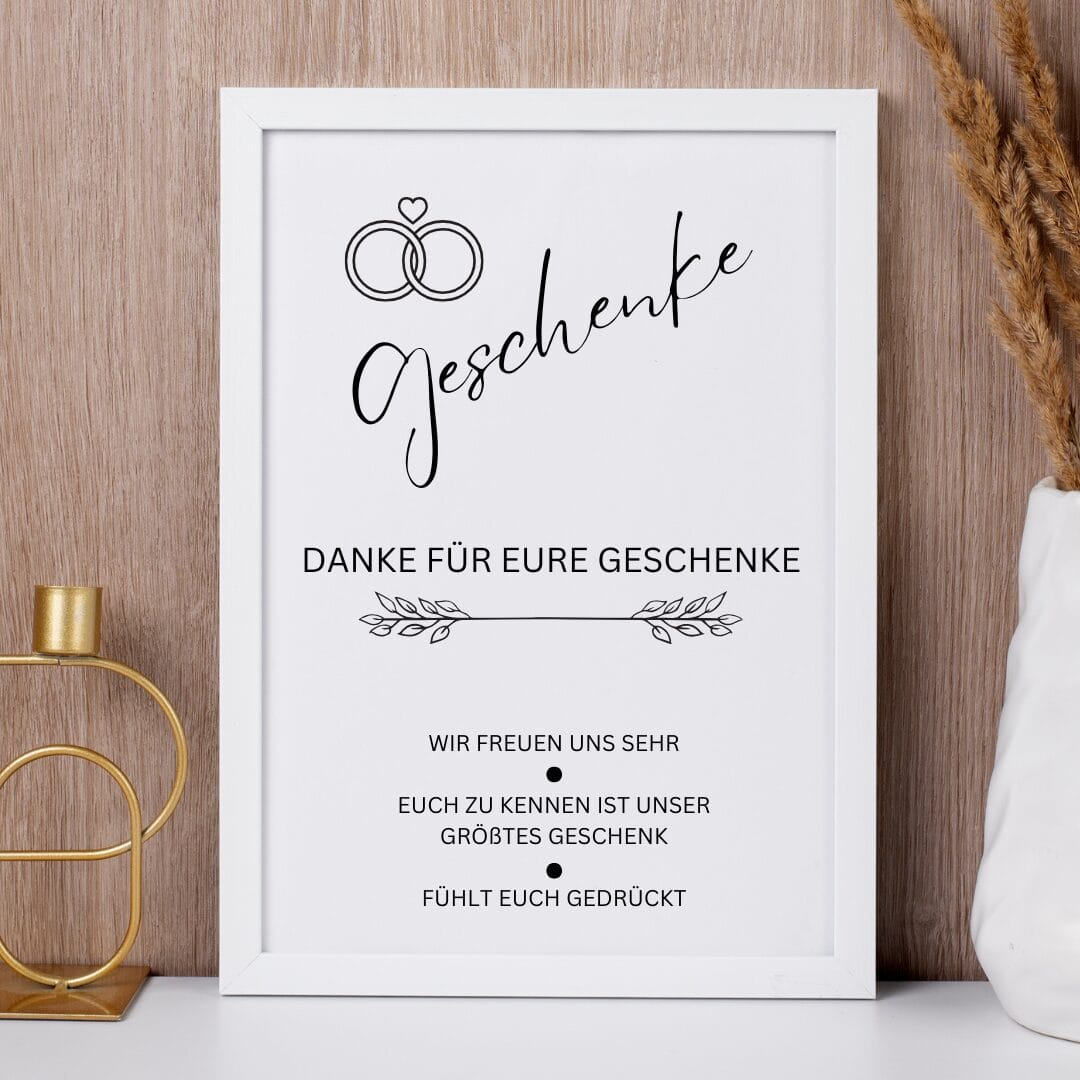 Ein gerahmtes Schild mit Eheringen und dem Wort Geschenke. Darunter ein deutscher Text, der den Gästen für ihre Geschenke dankt und sagt, dass es das größte Geschenk des Paares ist, sie zu kennen. Der Rahmen steht auf einer weißen Fläche mit dekorativen Elementen drum herum.