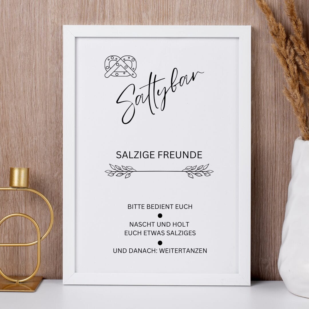 Ein gerahmtes Schild mit einer Brezelillustration und dem handgeschriebenen Text Saltybar. Darunter lädt ein deutscher Text die Gäste ein, sich an salzigen Snacks zu bedienen und dann weiterzutanzen. Der Rahmen steht auf einer Holzfläche neben einem goldenen Kerzenständer und getrocknetem Weizen.